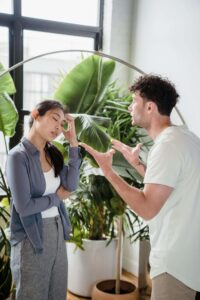 Couple in a room facing each other and communicating in a frustrated way. Male has hands out and female is resting one hand on head. 