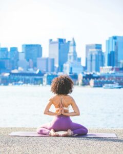 Person on yoga mat with view of the city in a seated pose with hand in prayer behind the back